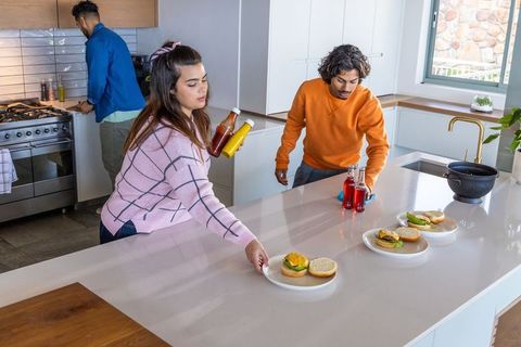 Friends preparing burgers on modern kitchen island holding condiments and beverages