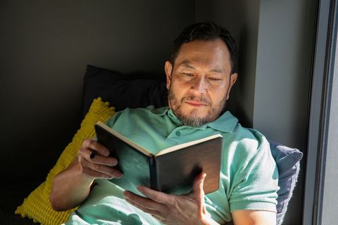 Mature asian man reading hardcover book in sunlit window nook, cozy minimalist interior
