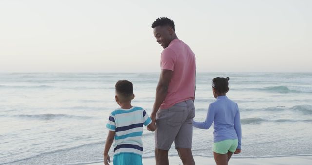 Father holding hands with his two children walking along shoreline during warm sunny day. Perfect for promoting family vacations, travel destinations, parenting topics, beach activities, and summer campaigns.