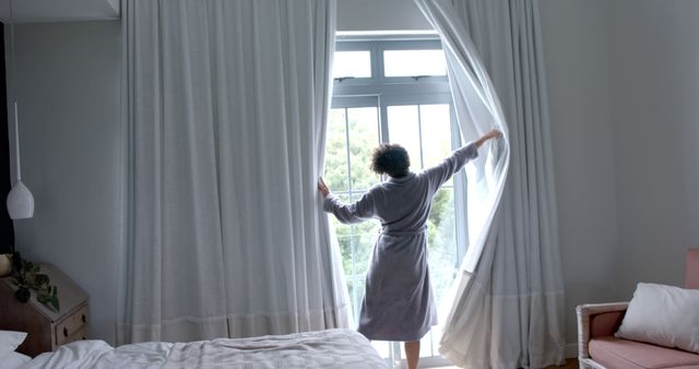 Woman dressed in cozy robe opening white curtains in bedroom to bright morning light. Ideal for themes associated with morning routines, waking up, starting day, and indoor lifestyle.