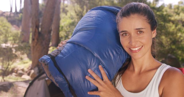 Cheerful woman carrying blue sleeping bag during camping trip in forest. Ideal for content about outdoor adventures, nature excursions, summer activities, camping gear reviews, and lifestyle blogs focused on active living and relaxation in nature.