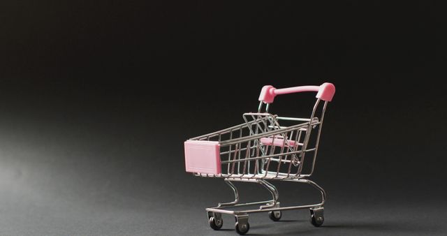 Miniature shopping cart with pink handles on dark background symbolizes retail, shopping, and commerce themes. Can be used for e-commerce websites, marketing material, business presentations, retail flyers, and creative advertisement campaigns.