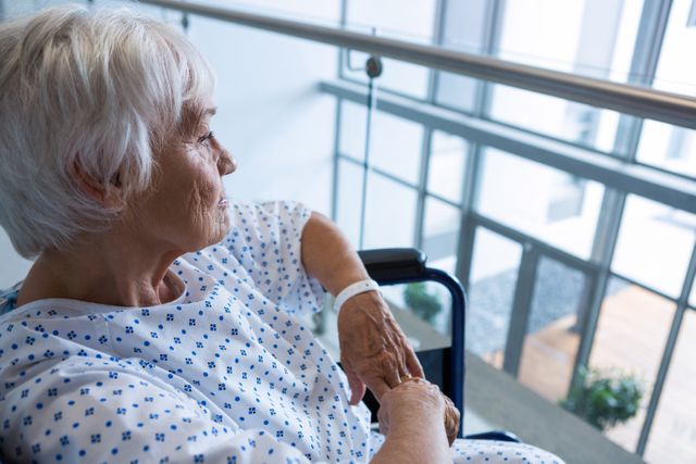 This image depicts an elderly patient in a wheelchair, wearing a hospital gown, looking thoughtful while sitting in a hospital passageway. It can be used in healthcare, medical, and senior care contexts to illustrate themes of patient care, recovery, and the challenges faced by the elderly in medical settings.