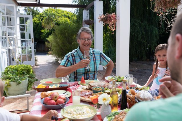 Family members of different generations are enjoying a meal together in a backyard setting. A man is toasting with a glass, indicating a celebratory or joyful moment. The table is filled with various dishes, fresh fruits, and drinks, suggesting a casual and relaxed atmosphere. This image can be used for themes related to family gatherings, outdoor dining, summer activities, and celebrating special occasions.