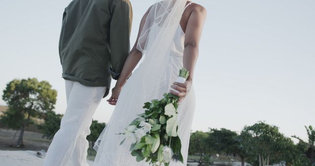 Midsection of biracial couple holding hands and flowers at wedding on beach. Love, wedding, ceremony, summer and lifestyle.