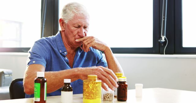 This image captures a senior man sitting at a table, thoughtfully organizing various medicine bottles. Ideal for illustrating topics related to elderly health care, medication management, aging, and senior citizen health awareness. It can be used in health-related articles, brochures, and websites specializing in elderly care or pharmaceutical services.