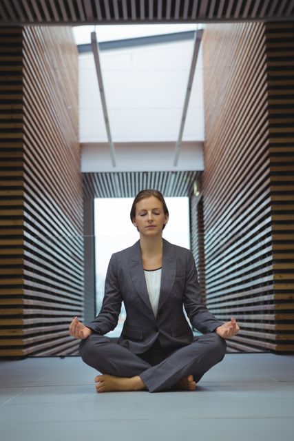 Businesswoman in a suit practicing yoga and meditation in an office corridor. Ideal for illustrating concepts of workplace wellness, stress management, and corporate mindfulness programs. Useful for articles, blogs, and promotional materials related to mental health, professional balance, and relaxation techniques in a corporate setting.