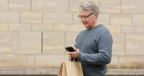 Mature man checking phone holding shopping bags by beige wall with glasses wearing sweater