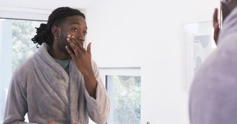 African American man applying moisturizer at mirror in gray bathrobe morning routine