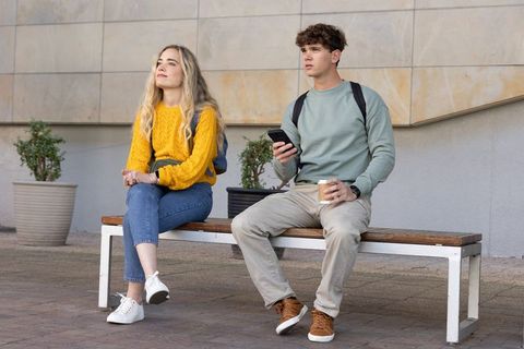 College students sitting on bench checking phone and sipping coffee on urban campus plaza