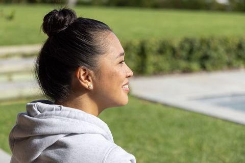 Young woman relaxing poolside on sunny lawn wearing gray hoodie and pearl stud earrings