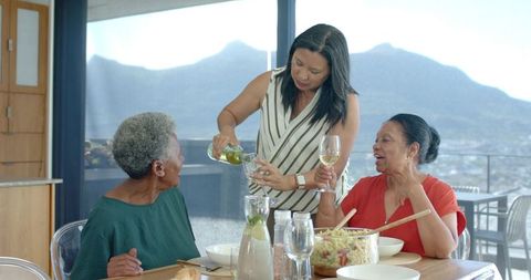 Senior african american and mature asian women pouring wine on balcony with mountain view