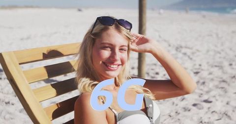 Blonde woman sunbathing on wooden deck chair at sunny sandy beach smiling with 6g overlay