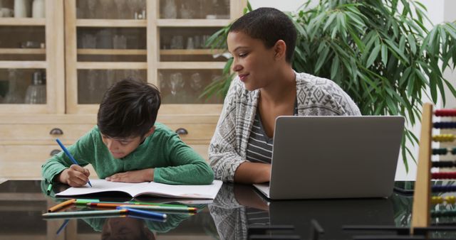 A mother is assisting her young son with his homework while working from home. They are at a kitchen table with school supplies and a laptop. This image is ideal for depicting the balance between family and remote work, online learning, homeschooling, and parental involvement in education.