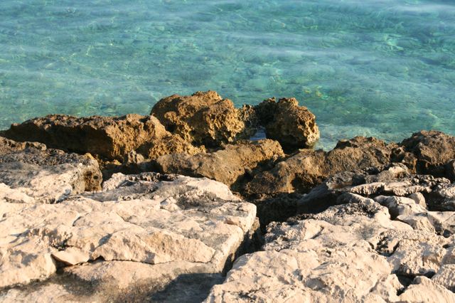 Rocky shoreline meeting crystal clear blue water on a bright sunny day. Ideal for travel brochures, nature websites, coastal promotions, beach holiday themes, and serene landscape displays. Perfect for illustrating natural beauty, tranquility, and summer vacation destinations.