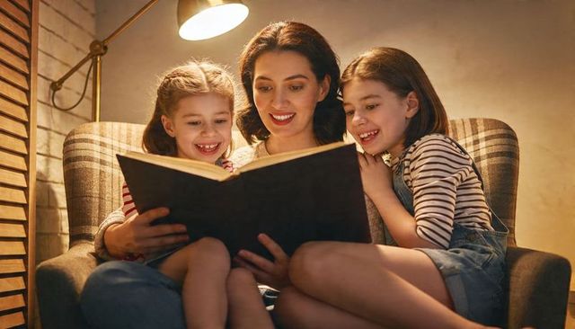Mother reading bedtime stories with two smiling daughters in cozy armchair under warm lamp