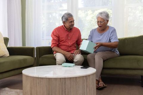 Senior couple opening teal gift on green sofa, surprised woman smiling, cozy living room moment