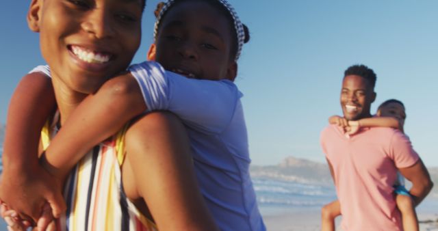 Happy African American family spending time together at a beach. Mother and daughter showing joyful expressions, while father and son enjoying their day near water with a backdrop of blue sky and ocean waves. Ideal for use in content related to family activities, vacations, summer fun, outdoor bonding, and healthy lifestyle.