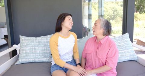 Asian mother and daughter sitting on porch sofa holding hands sharing warm conversation