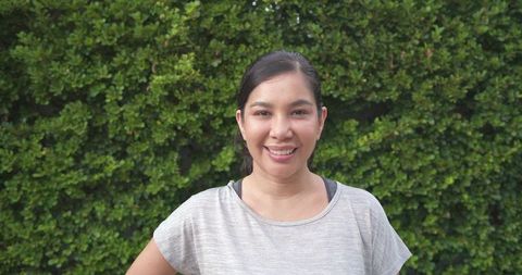 Smiling mid-adult woman standing against lush green hedge wearing gray top and visible bra straps