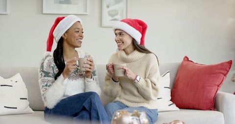 Multicultural women wearing santa hats sharing cozy holiday mugs and chatting on sofa