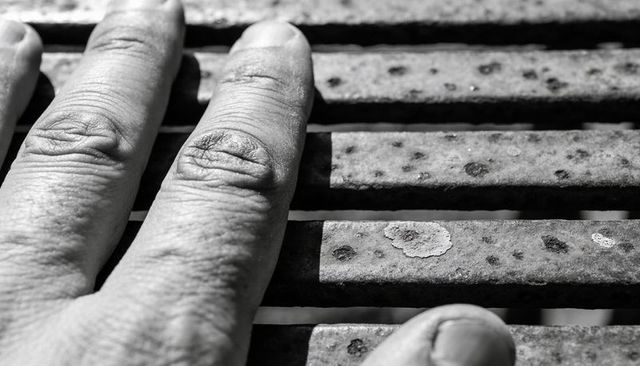 Left hand resting on rusted metal bench slats close-up monochrome texture detail gritty