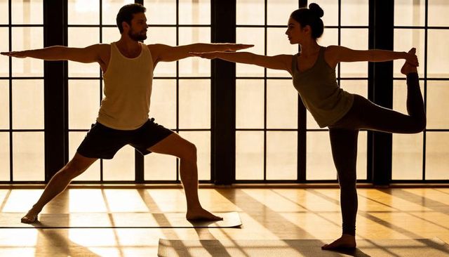 Couple practicing yoga in sunlit studio balancing warrior and dancer poses in silhouette