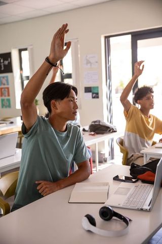 Students raising hands in bright classroom with laptop notebook and headphones