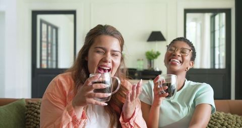 Diverse friends laughing holding double-walled glass coffee mugs on cozy living room sofa