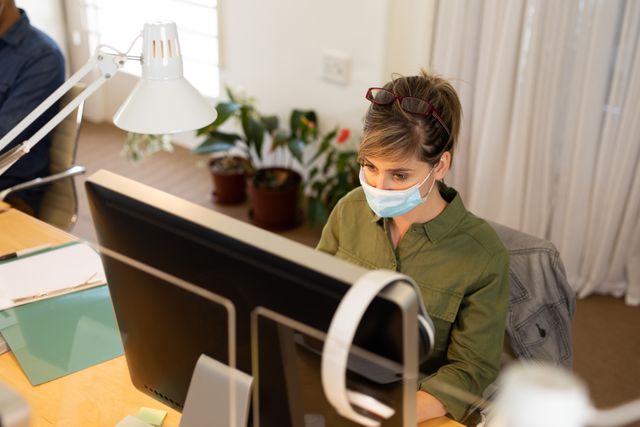 Caucasian woman wearing a facemask working at her desk in an office environment. She has her glasses pulled up on her head and is focused on the computer screen. This image can be used to depict workplace safety measures during the pandemic, professional work settings, and health precautions in business environments.