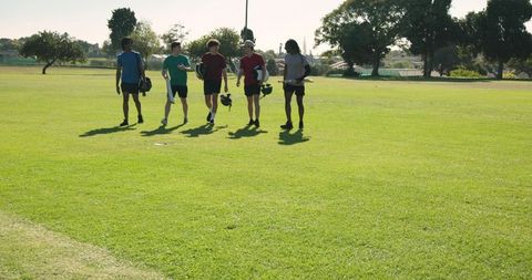 Young diverse cricket teammates walking across sunlit field carrying helmets and boards