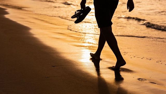 Sunset silhouette of barefoot man walking along wet shoreline holding sandals leaving footprints