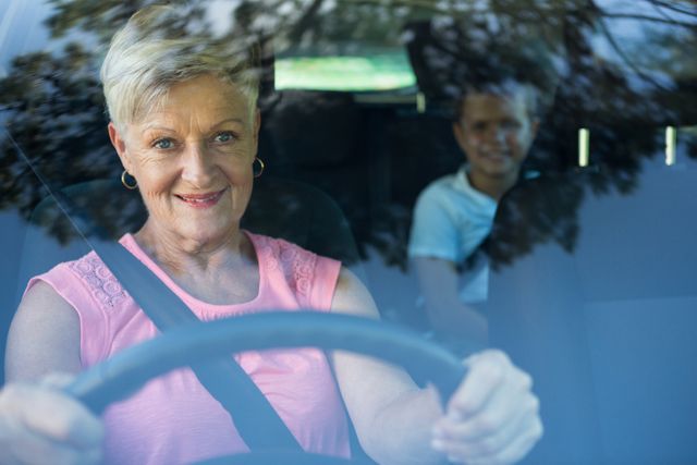 Senior woman driving car with young grandson sitting in the back seat. Both are smiling and enjoying the ride. Ideal for use in family travel, road safety, and intergenerational bonding themes.