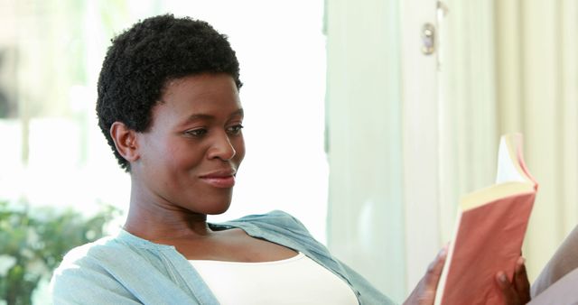 This image shows an African American woman reading a paperback book while sitting at home, appearing relaxed and content. Ideal for use in articles or blogs on leisure activities, home lifestyle, literature, or personal relaxation techniques. Suitable for promoting comfortable furniture, reading material, or home decor.