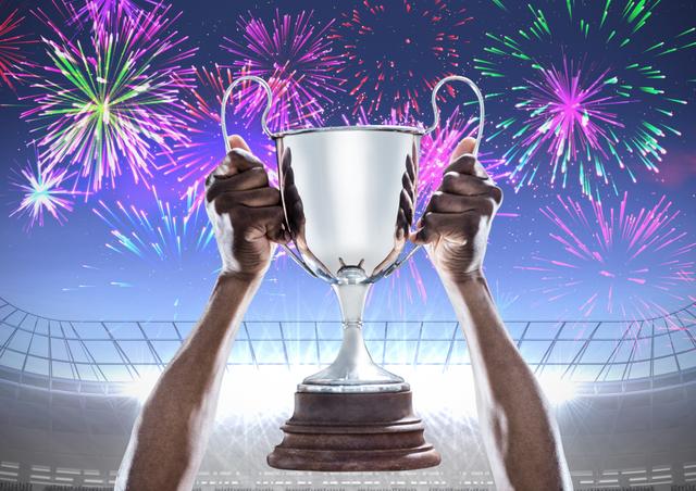 Hands holding a trophy with fireworks in the background, set in a stadium. Ideal for use in sports-related promotions, victory celebrations, motivational posters, and achievement-themed content.