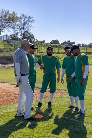 Coach leading baseball drill, players gathering around cone on sunny grass and dirt field