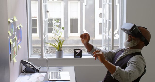 Senior businessman wearing a VR headset while analyzing data displayed on the wall of a modern office. Desk includes laptop, charts, flower pot, and smartphone. Ideal for illustrating innovative business solutions, modern technology integration in business, remote work, and senior professionals adapting to new tech advancements.