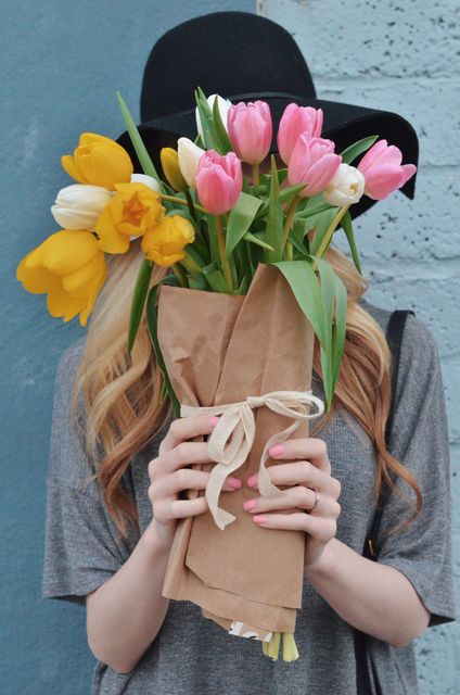 Woman holding a mixed bouquet of pink tulips and yellow daffodils wrapped in brown paper. Hands are visible with well-groomed nails, and she is wearing a black hat and grey top. Perfect for use in floral arrangement promotions, gift ideas, or spring season advertisements.