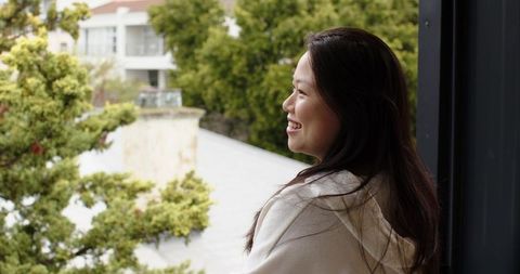 Asian woman standing on balcony smiling while overlooking calm urban neighborhood