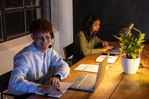 Evening customer support team collaborating on laptops with headsets, notebooks and charts