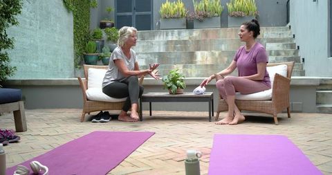 Two senior women sitting and talking on patio after yoga, wearing activewear and smiling