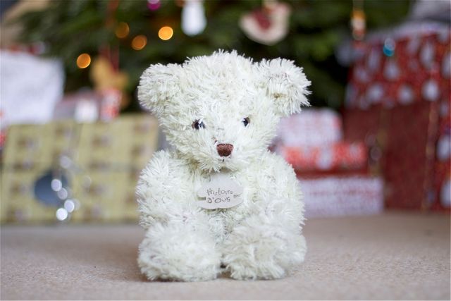 Fluffy teddy bear sitting in front of a Christmas tree with colorful gifts in the background. Ideal for holiday season promotions, greeting cards, and family-oriented advertisements. Can also be used in children's content or online stores selling toys and gifts.