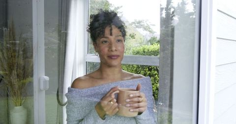 Mid-adult woman holding mug, gazing through window at garden, cozy off-shoulder sweater moment