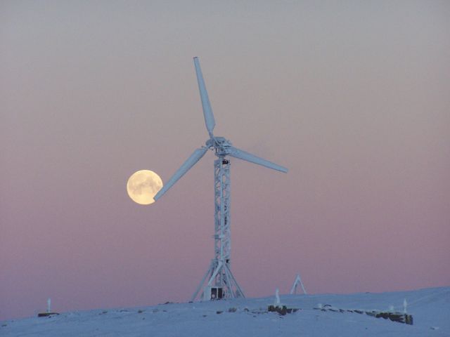 This serene winter scene depicts a full moon rising behind a wind turbine set in a snowy landscape under a pink sunset sky. Ideal for illustrating themes related to renewable energy, clean technology, and the peacefulness of nature during winter evenings.