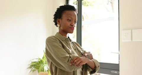 Contemplative african american woman standing by glass door with arms crossed in bright home