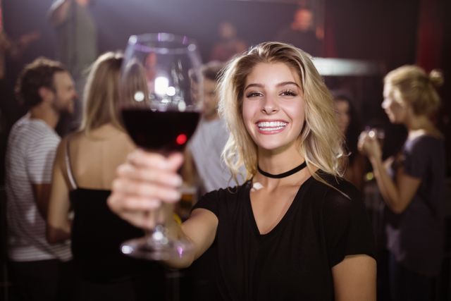 Young woman enjoying a night out at a nightclub, holding a wineglass and smiling at the camera. Ideal for use in advertisements for nightlife venues, social events, or promotional materials for bars and clubs. Can also be used in lifestyle blogs or articles about socializing and nightlife.