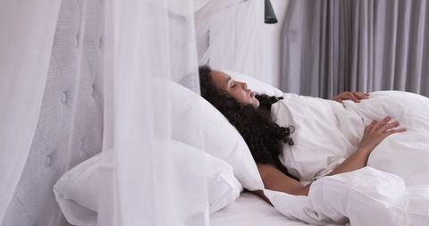 African american woman resting on white duvet under sheer canopy, serene bedroom morning