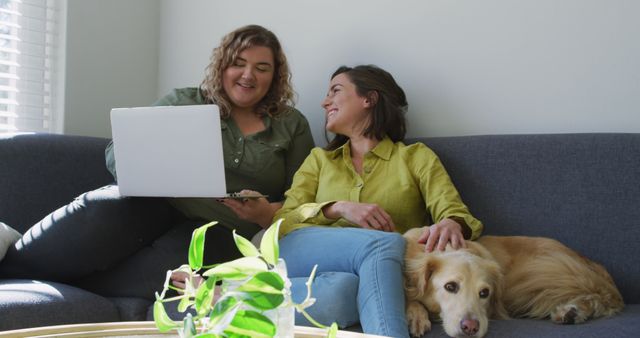 Caucasian lesbian couple using laptop and sitting on couch with dog. domestic life, spending free time relaxing at home.