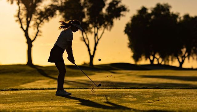 Swinging teenage female golfer striking ball on fairway at golden hour sunset silhouette