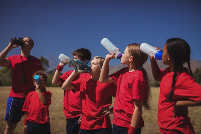 Group of children and their trainer drinking water during an outdoor boot camp on a sunny day. Ideal for use in content related to fitness, outdoor activities, children's health, teamwork, and summer camps.